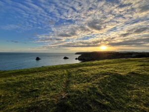 A landsca[e view of a field in the foreground, dropping off into the background of sea and cliffs against a setting sun.
