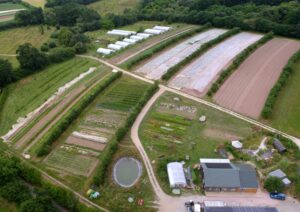 Green fields showing a regenerative farm from a birds-eye view. Some of the crop fields are covered in netting, there is a farm building in the lower right corner, and polytunnels in the upper left corner.