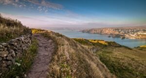 A path snaking to the left, with soft green grass sloping down the hill, with a view of the sea and distant cliff faces