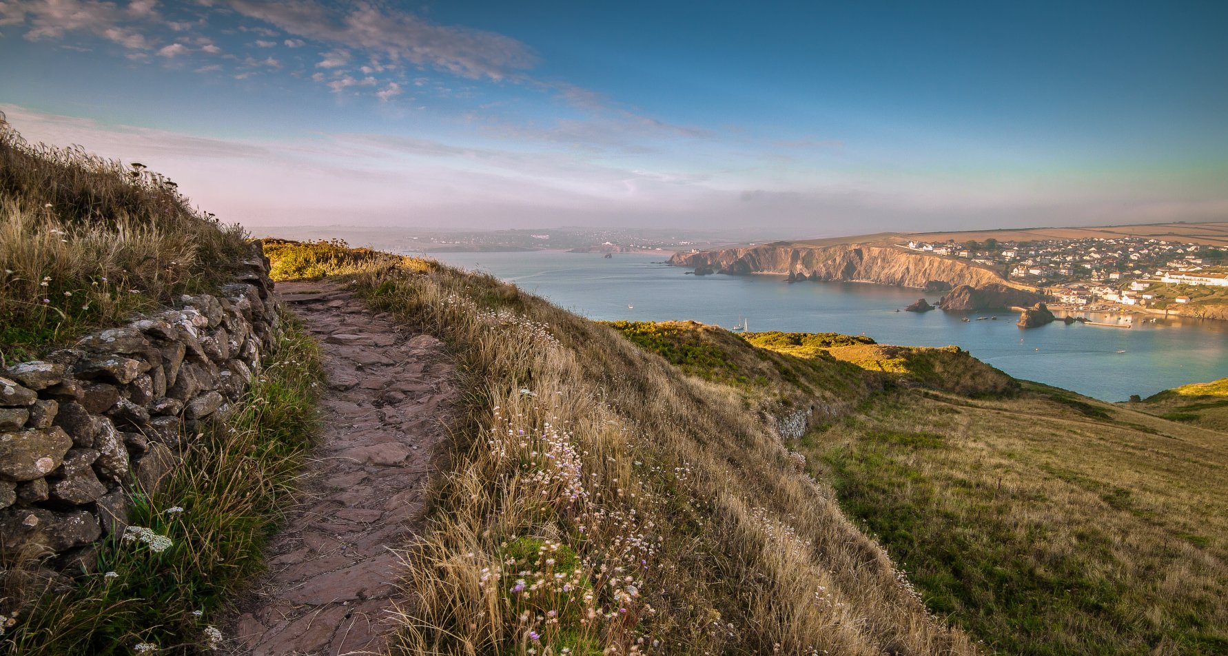 A path snaking to the left, with soft green grass sloping down the hill, with a view of the sea and distant cliff faces