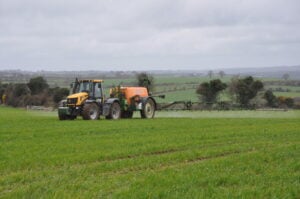 A yellow, orange and green tractor sprays a green field full of crops with fields, trees, and hedgerows in the background.
