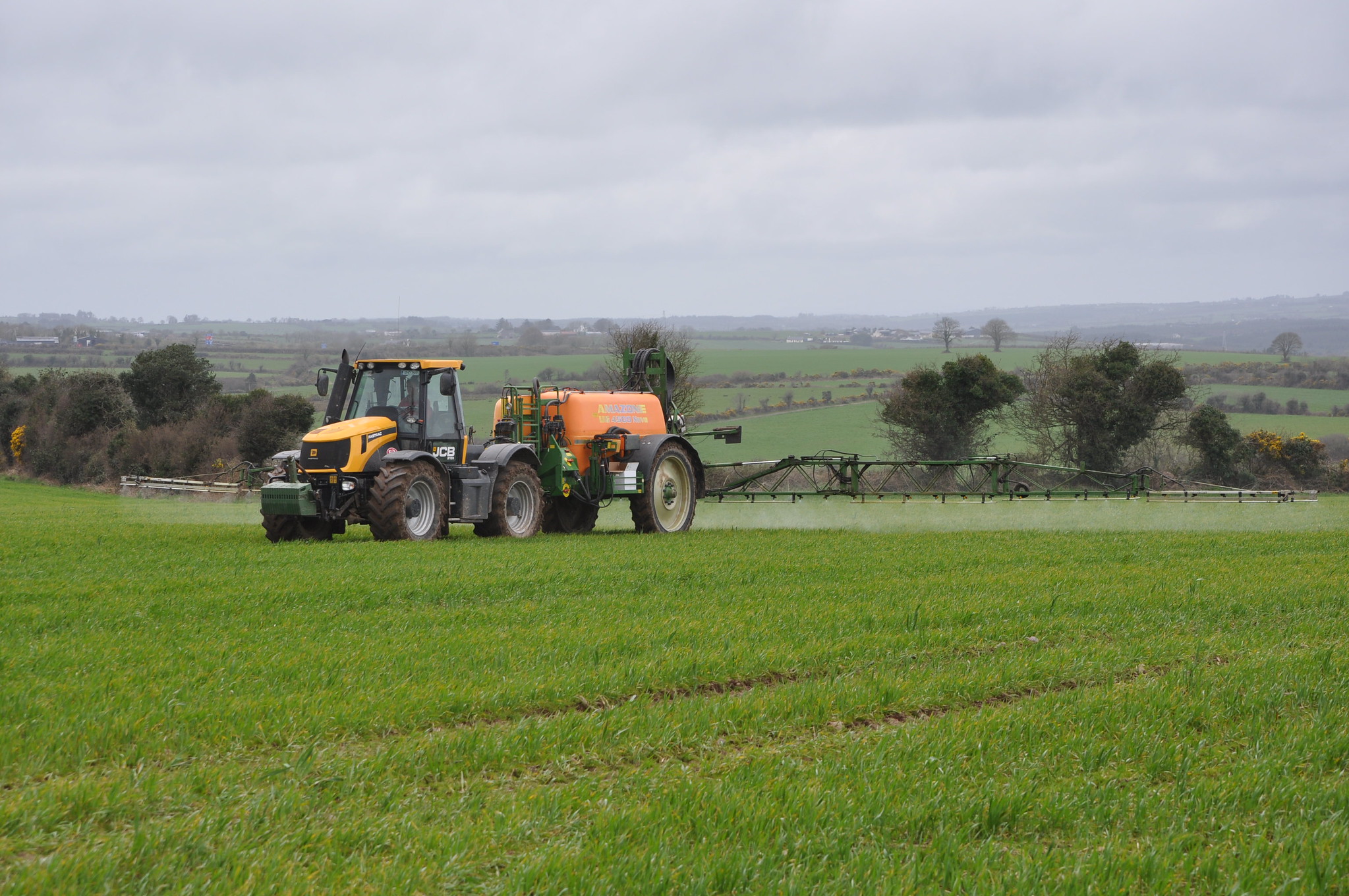 A yellow, orange and green tractor sprays a green field full of crops with fields, trees, and hedgerows in the background.